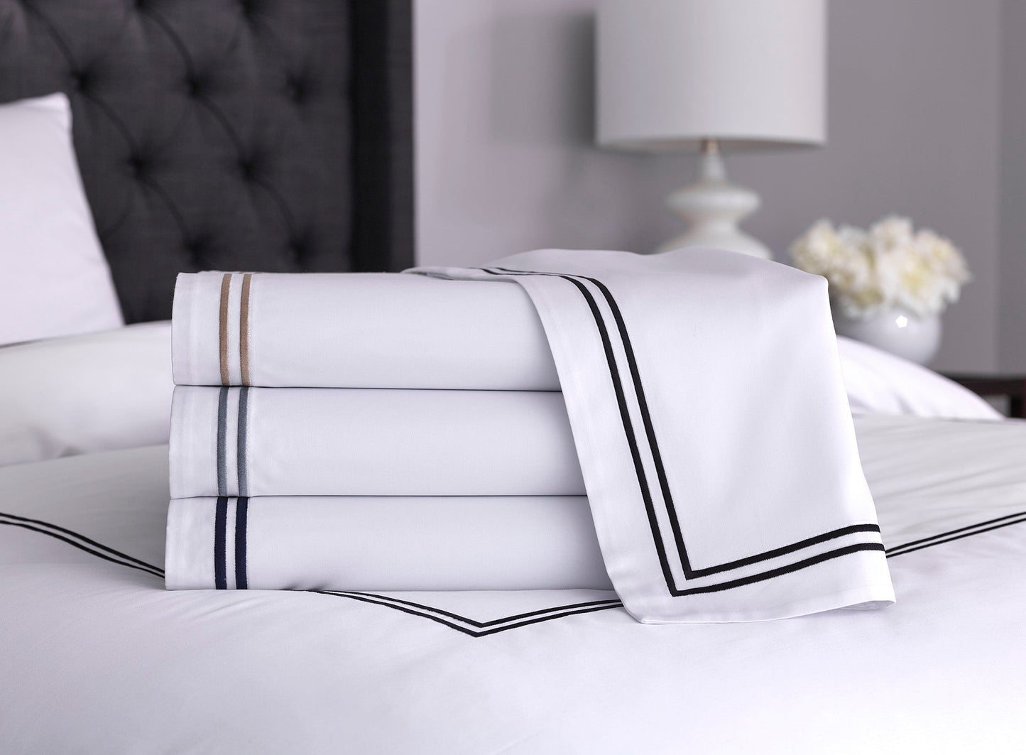 Front view of folded white cotton sheets with black, silver and warm taupe stitching, highlighting their soft and silky texture and clean finish 